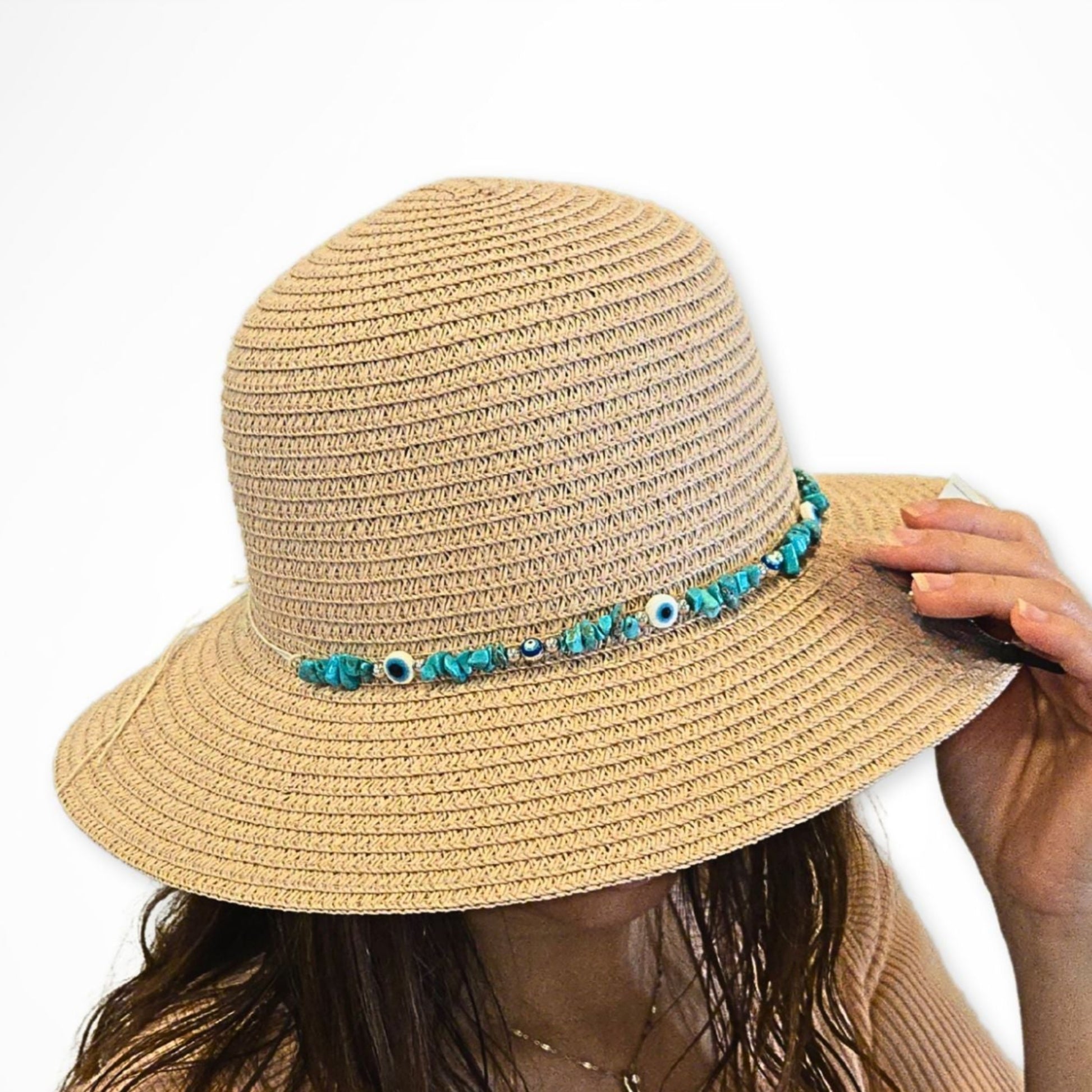 A straw hat with a beige color and a charm featuring an evil eye and beads around the crown, worn by a person with their hand adjusting the brim.