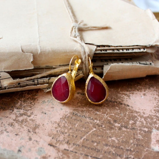 A pair of teardrop-shaped earrings with red carnelian stones set in gold-plated sterling silver, displayed on a textured background with a book and paper.