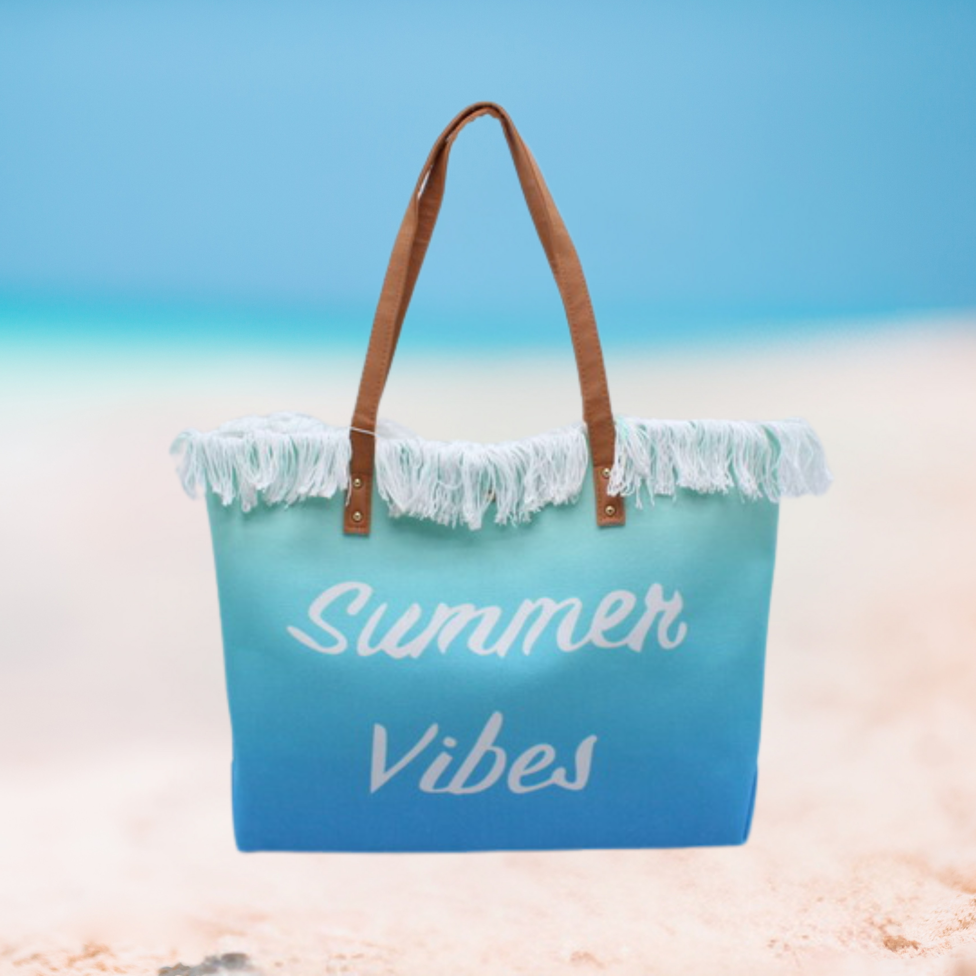 A beach tote bag with a blue gradient design, white fringe trim, and brown handles, featuring the words 'Summer Vibes' in white script, set against a blurred beach background.