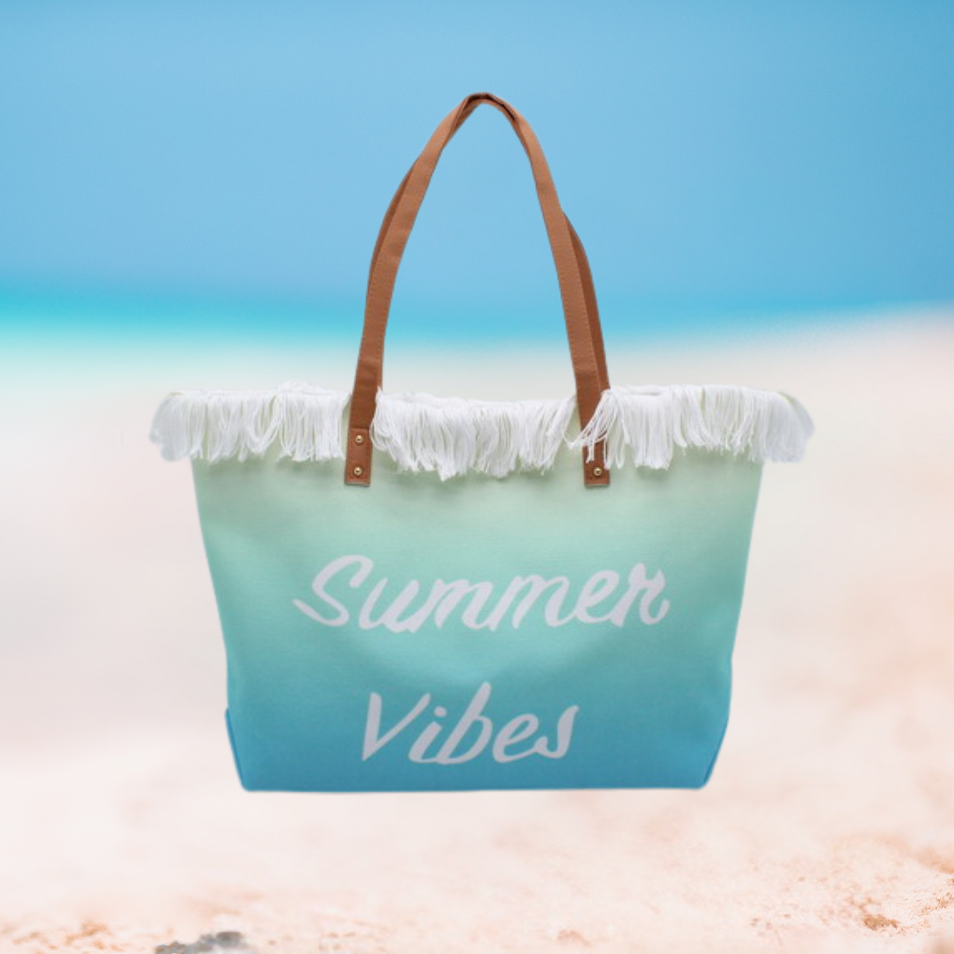 A beach tote bag with a blue-to-teal gradient, white fringe trim, and brown handles, featuring the words 'Summer Vibes' in white script, set against a blurred beach background.