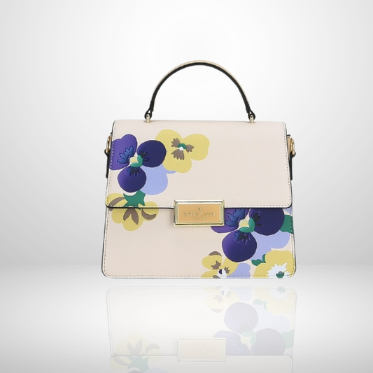 A structured top-handle handbag in light blush pink with a floral print of purple, yellow, and blue pansies. The bag features a gold-tone metal brand plaque on the front flap and a black top handle, set against a clean white background with a soft reflection below.