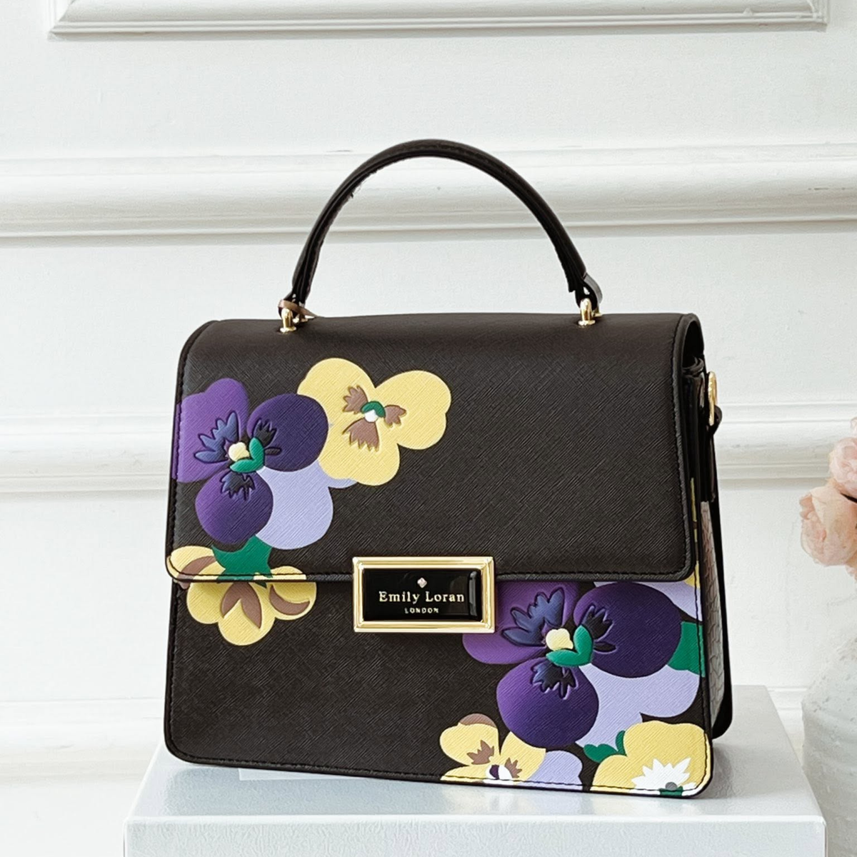 A black textured handbag with a top handle, featuring a colorful floral print of purple, yellow, and lavender pansies on the front and bottom flap. It has a gold-tone metal brand plaque reading 'Emily Loran London' and is displayed on a white pedestal against a white paneled wall.