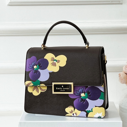 A black textured handbag with a top handle, featuring a colorful floral print of purple, yellow, and lavender pansies on the front and bottom flap. It has a gold-tone metal brand plaque reading 'Emily Loran London' and is displayed on a white pedestal against a white paneled wall.