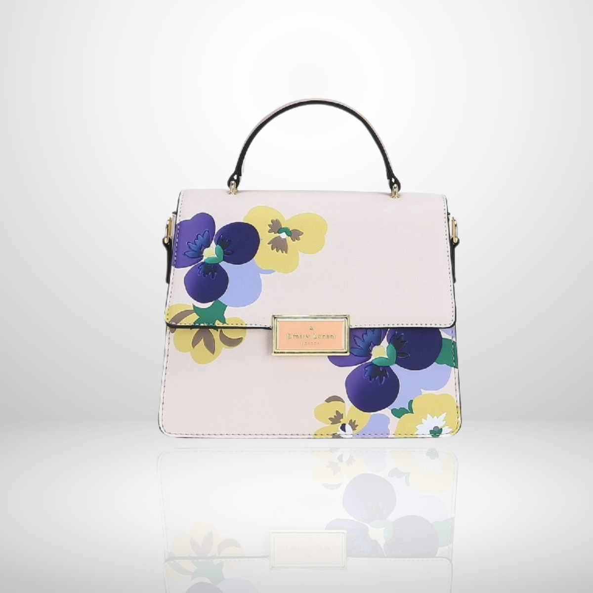 A structured top-handle handbag in pale blush pink with a colorful floral print of purple, yellow, and blue pansies. It features a gold-tone metal plaque on the front flap and a black top handle, set against a clean white background with a soft reflection below.