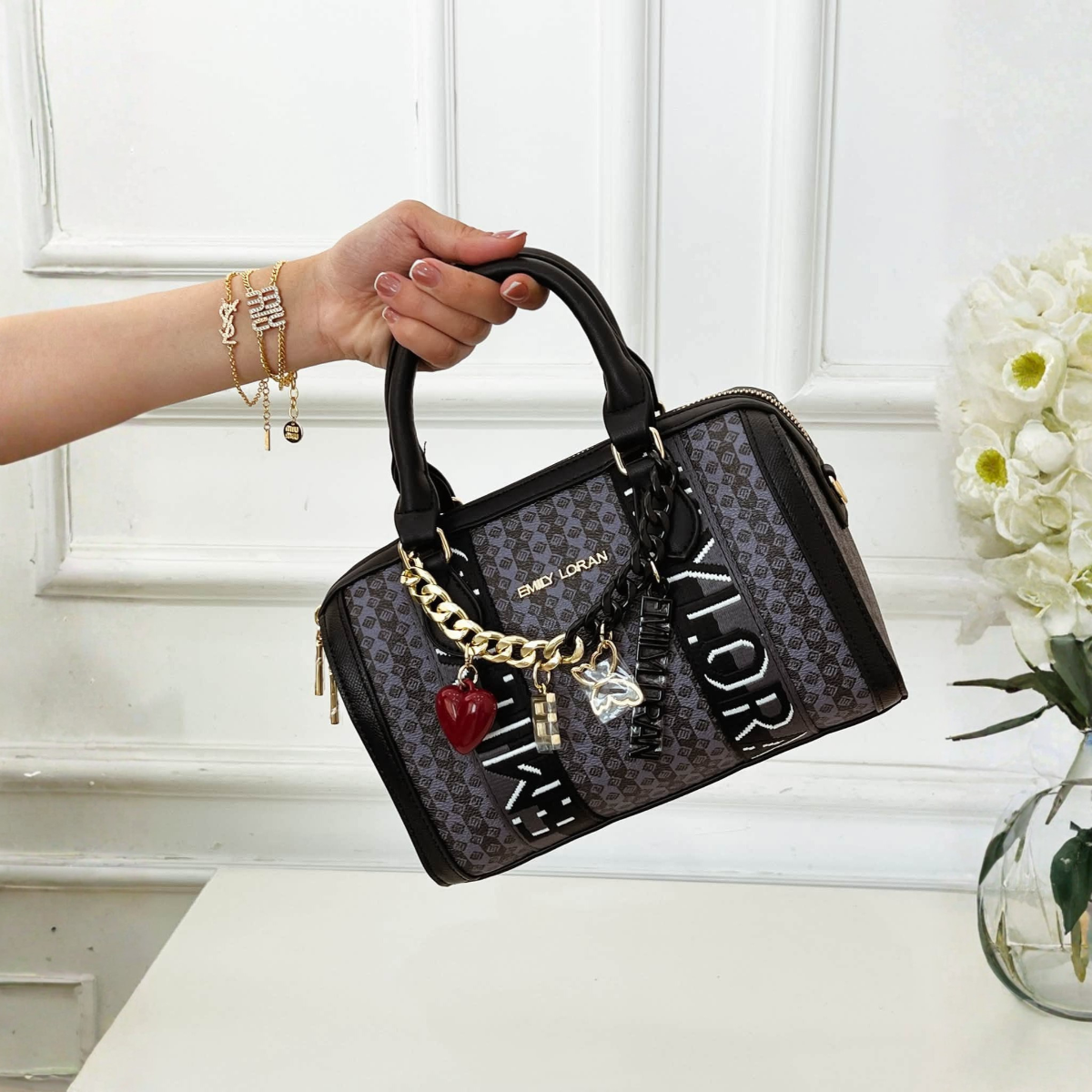 A hand holds a barrel handbag with a patterned body, black leather trim, and gold-tone chain charms, set against a white paneled wall and flowers
