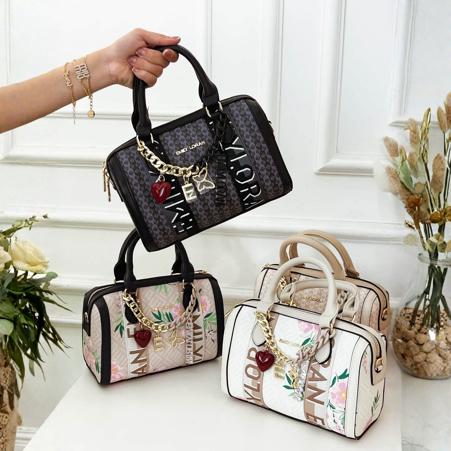 A hand holds a barrel handbag with a patterned body, black leather trim, and gold-tone chain charms, set against a white paneled wall and flowers