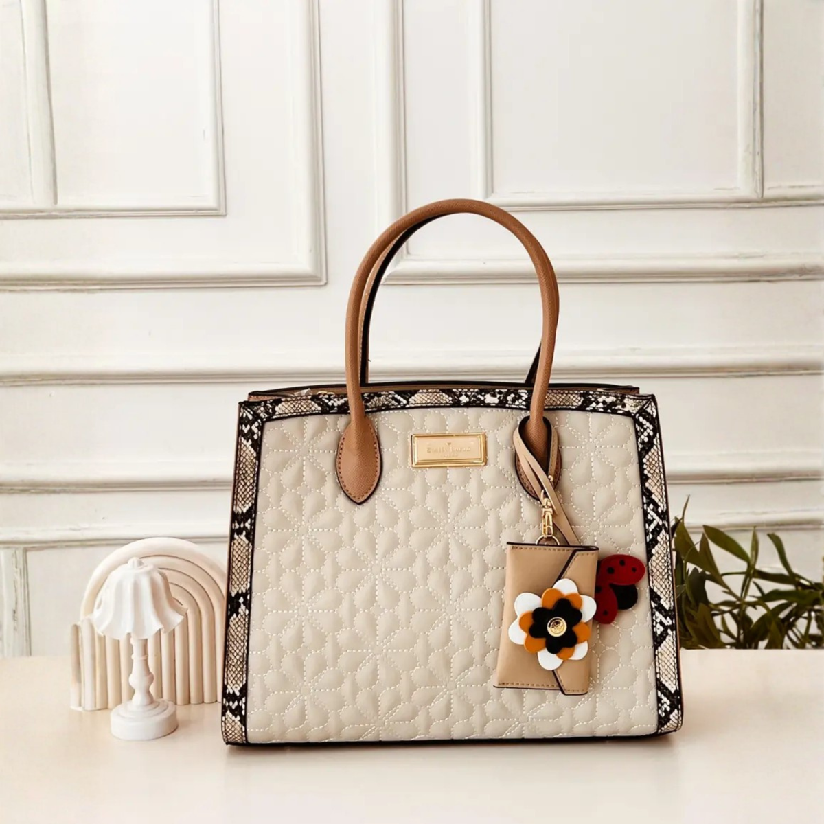 A structured tote bag with a cream quilted front, black and beige snakeskin-patterned trim, and tan leather handles, styled on a tabletop with a small decorative lamp and greenery in the background.