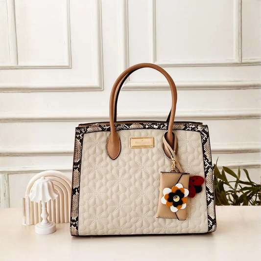 A structured tote bag with a cream quilted front, black and beige snakeskin-patterned trim, and tan leather handles, styled on a tabletop with a small decorative lamp and greenery in the background.