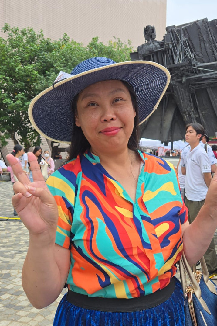 Woman in colorful shirt and blue hat making peace signs outdoors